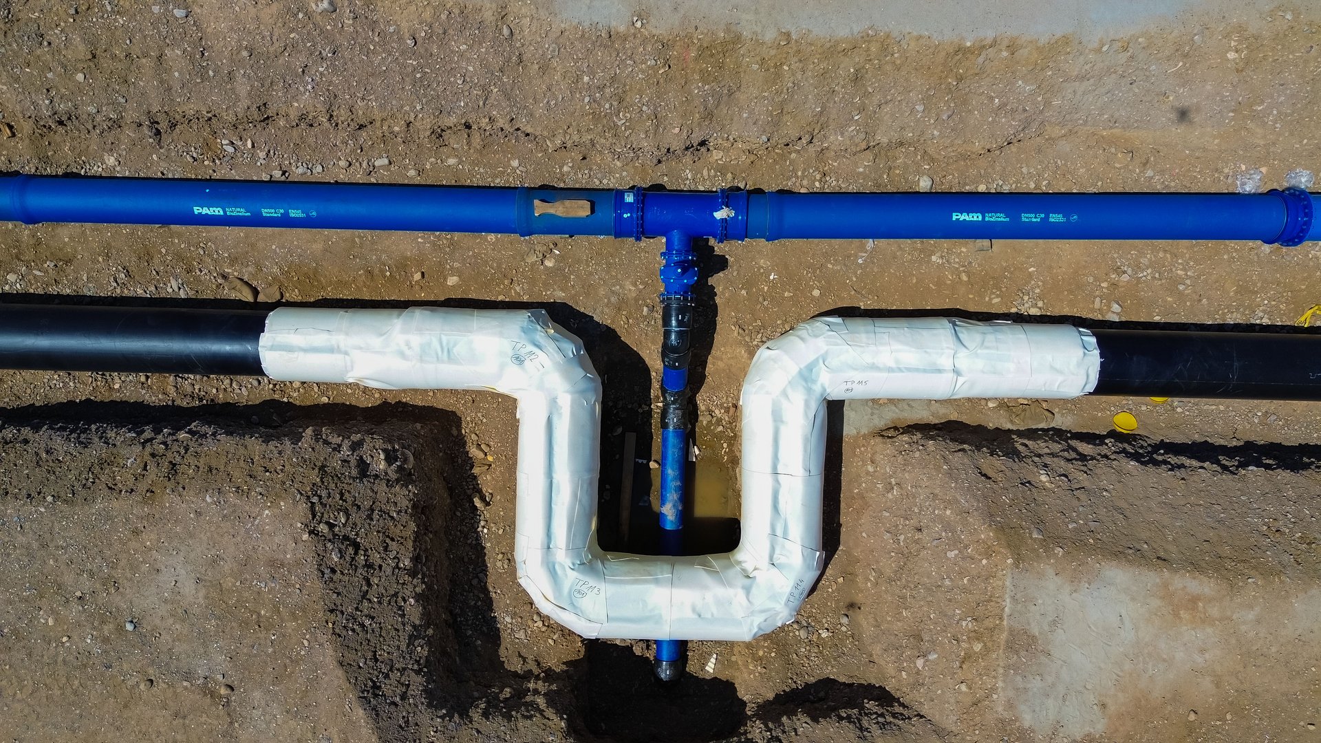 Eine Konstruktion, bei der sich blaue Trinkwasser- und schwarze Fernwärmerohre überkreuzen. Die Rohre sind bereits mit weißem Schaumstoff isoliert.