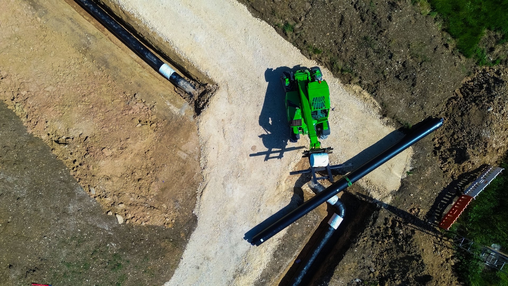 Eine Baustelle mit einem großen grünen Teleskoplader, der ein schwarzes Fernwärmerohr anhebt. Mehrere schwarze Rohre liegen in einer ausgehobenen Grube.